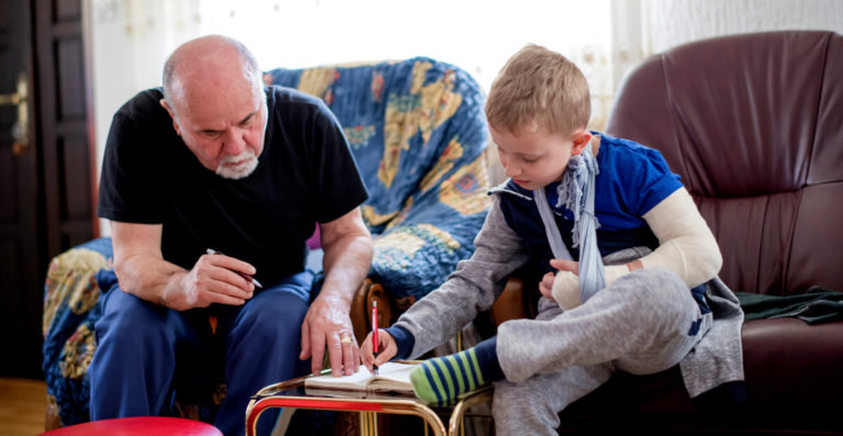 A grandparent helps a school-aged child write in a journal. The child has a bandaged and slinged arm, and is writing in the journal with their unaffected hand.