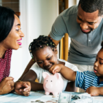 Two parents and two young children sit at a table, with a piggy bank between them all. One of the children is adding a coin to the piggy bank, with the parents smiling and looking on. On the table, there are coins and bills of various denominations.