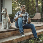A mature man at his cottage resting on the porch with his dog. Sitting in a chair, drinking coffee, and using a smartphone.