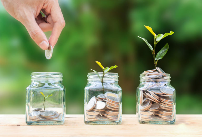 A man hand holding a coin over stacked coins in a glass jar and growing trees planted from the coins as a metaphor for financial growth.