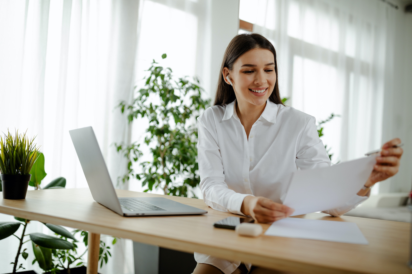 Smiling young Caucasian woman holding paper bill letter doing paperwork bill reading good news, checking post mail sitting at home table. Happy lady customer receive bank receipt sheet tax refund notification, wearing wireless headphones.