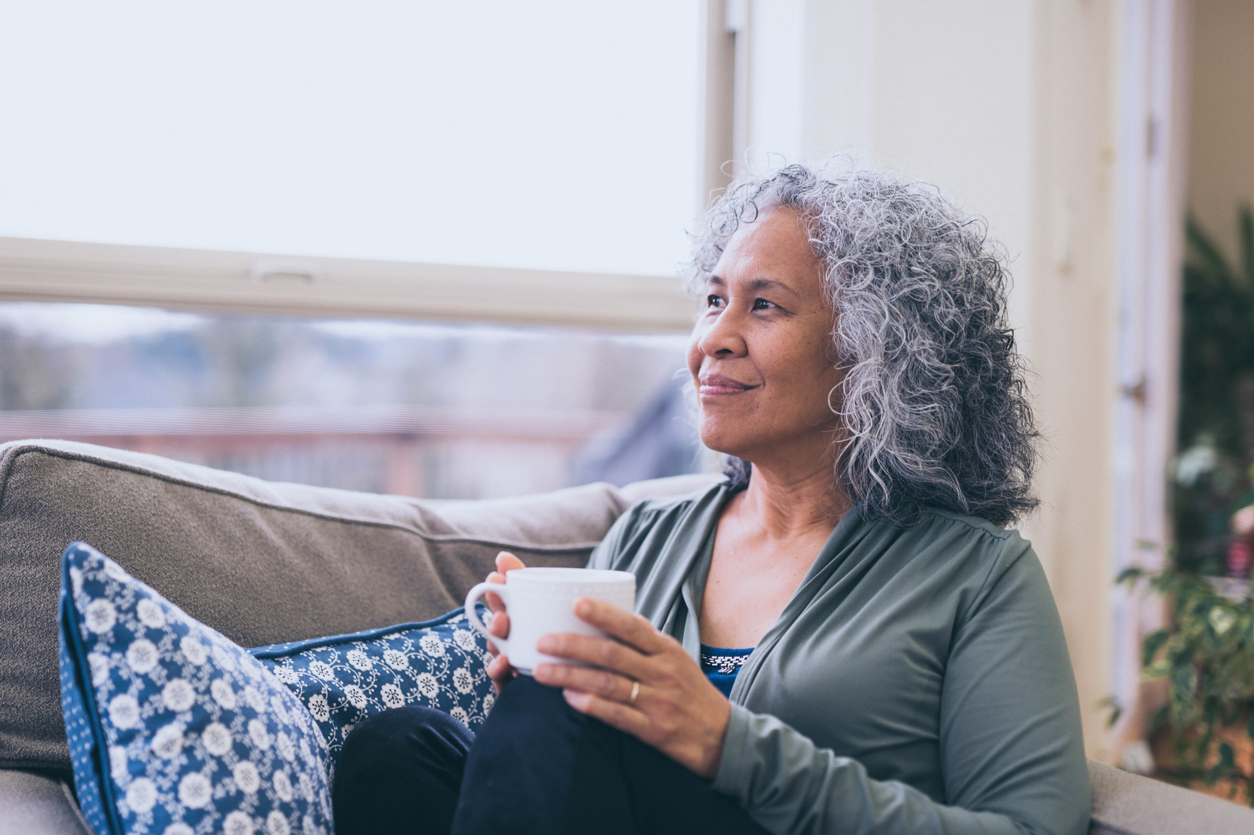 Mature woman sitting with her coffee in a comfy chair, looking out the window, smiling. 