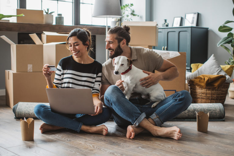 A happy young couple just moved into a new house. They are sitting on the floor with their dog, using a laptop to buy stuff they need.